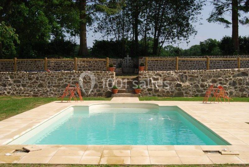 Piscine extérieure rectangulaire entourée de dalles claires, avec un mur en pierre et des arbres en arrière-plan.