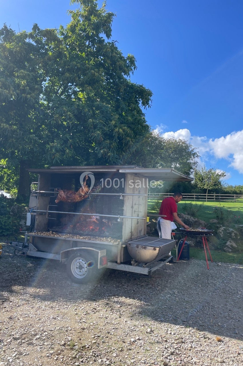 Un cuisinier prépare un cochon rôti sur une broche dans un espace extérieur ensoleillé, entouré de verdure.