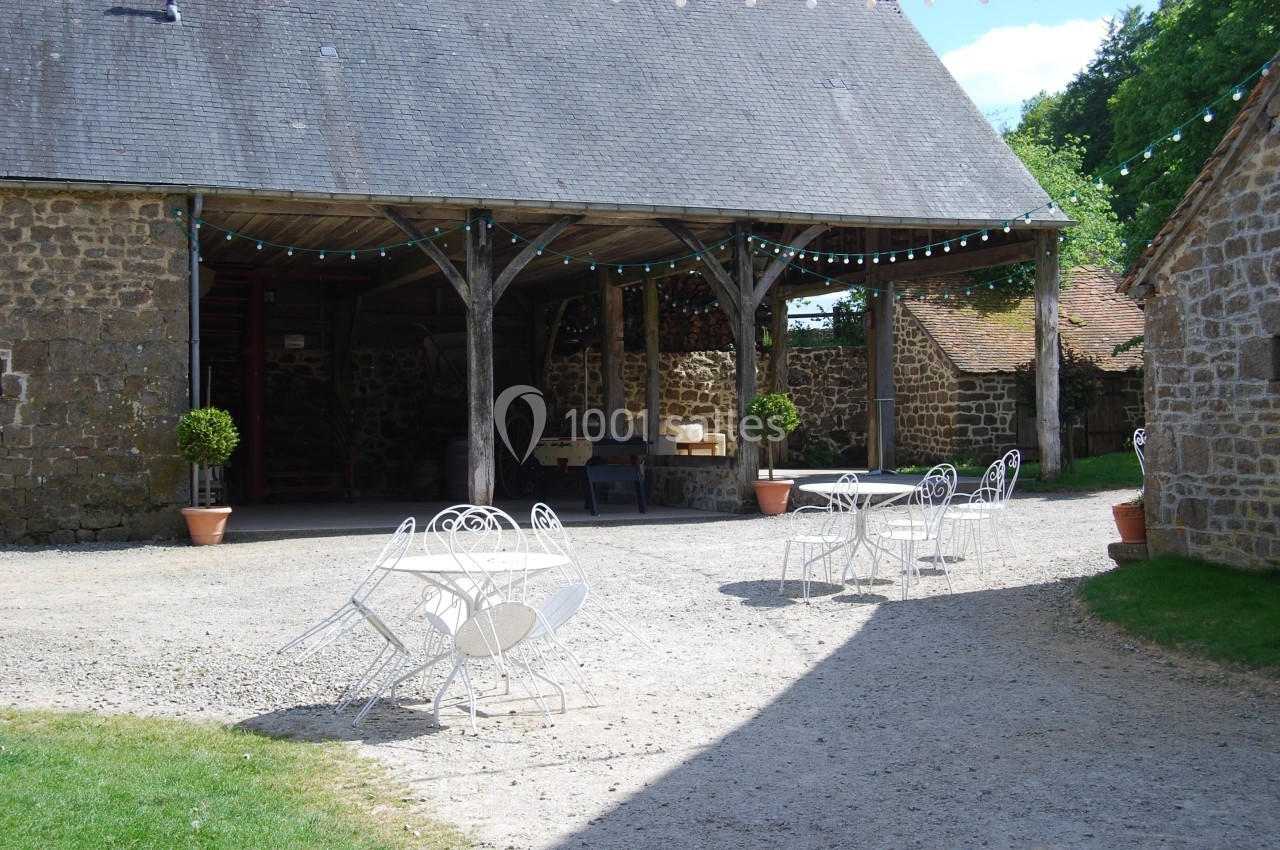 Cour d'une ferme avec une structure en pierre et bois, tables blanches en fer forgé et guirlandes lumineuses suspendues.