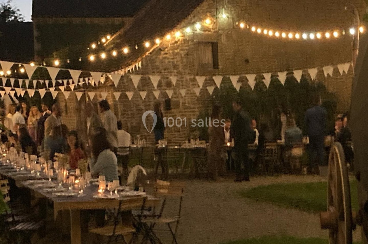 Cour extérieure éclairée par des guirlandes lumineuses, avec des tables dressées et des invités rassemblés en soirée.
