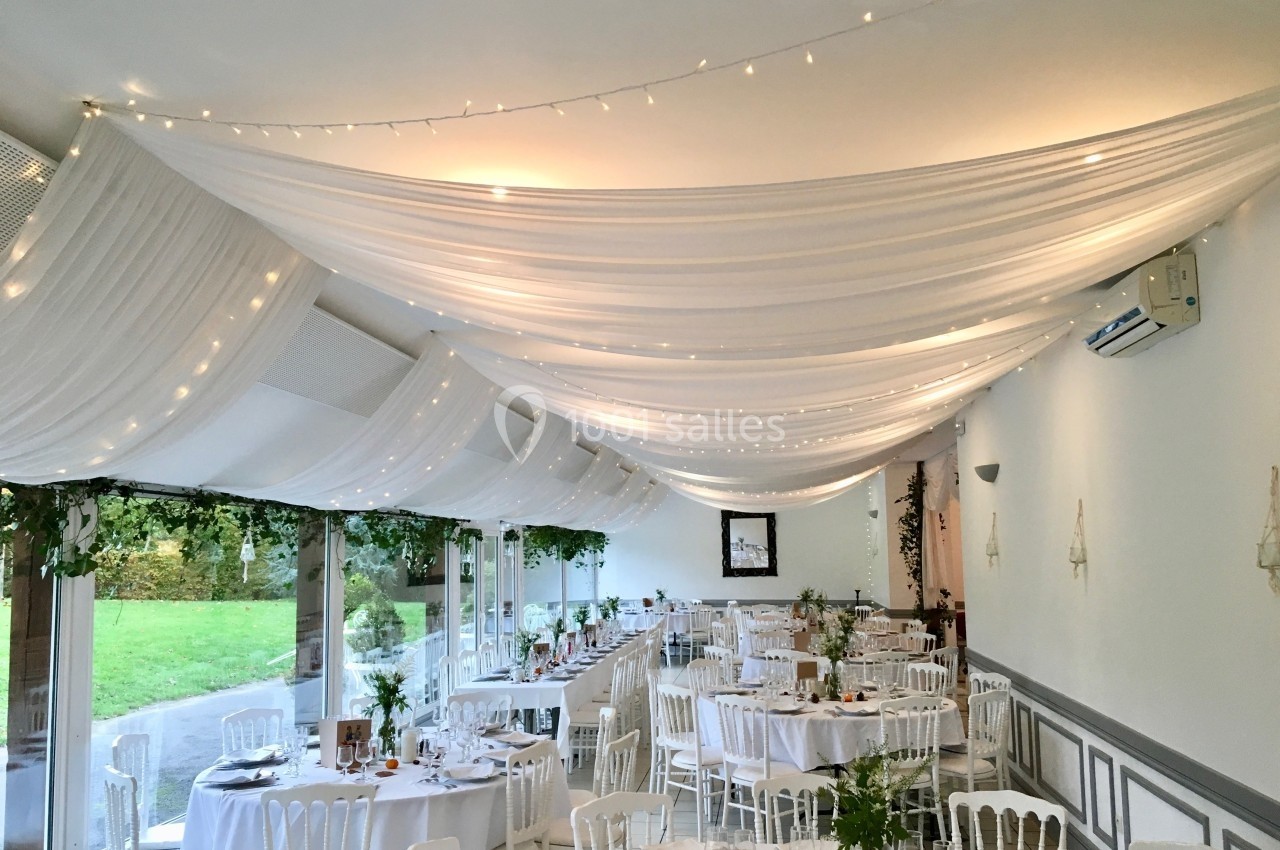 Salle de réception décorée avec des drapés blancs au plafond, des guirlandes lumineuses et des tables dressées.