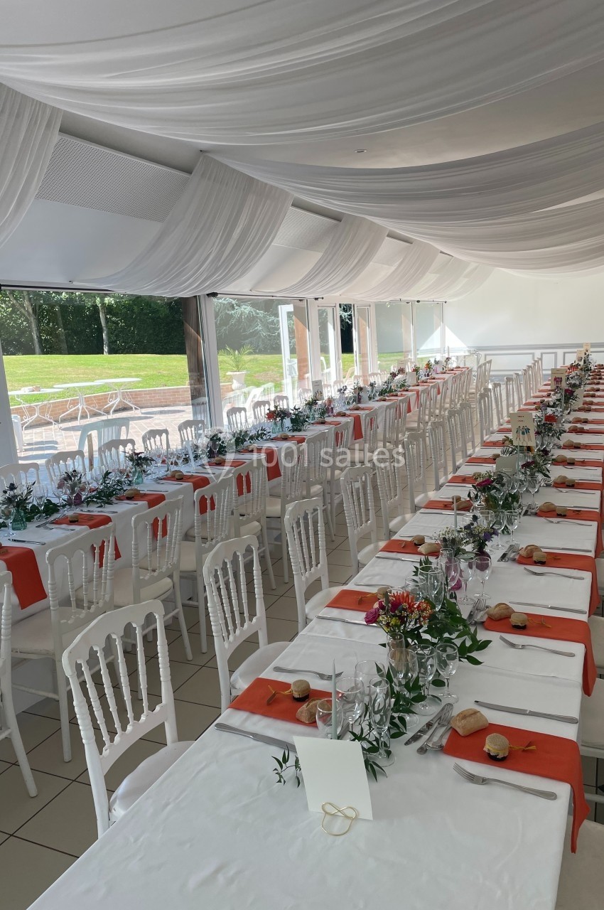 Salle de réception décorée avec des tables alignées, nappes blanches, chemins de table orange et chaises blanches.