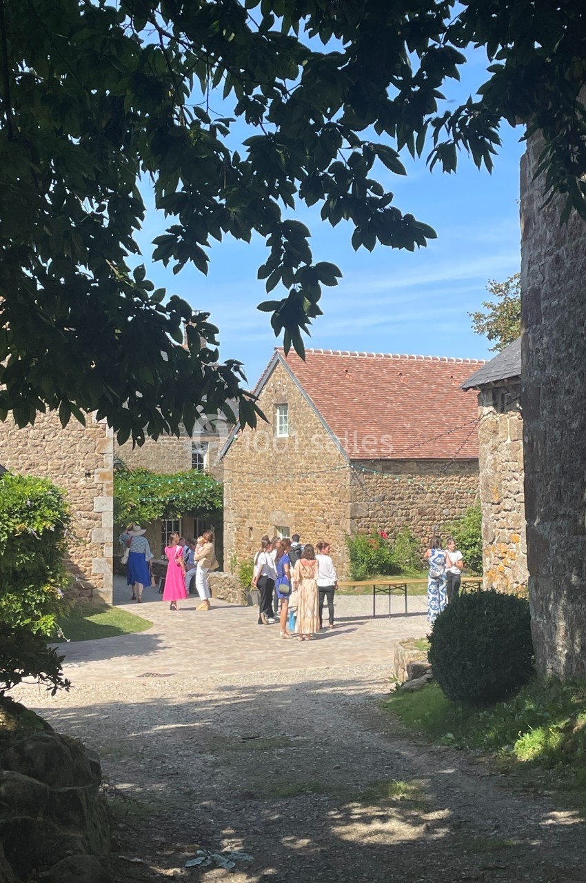 Un groupe de personnes se tient dans une cour pavée entourée de bâtiments en pierre sous un ciel dégagé.