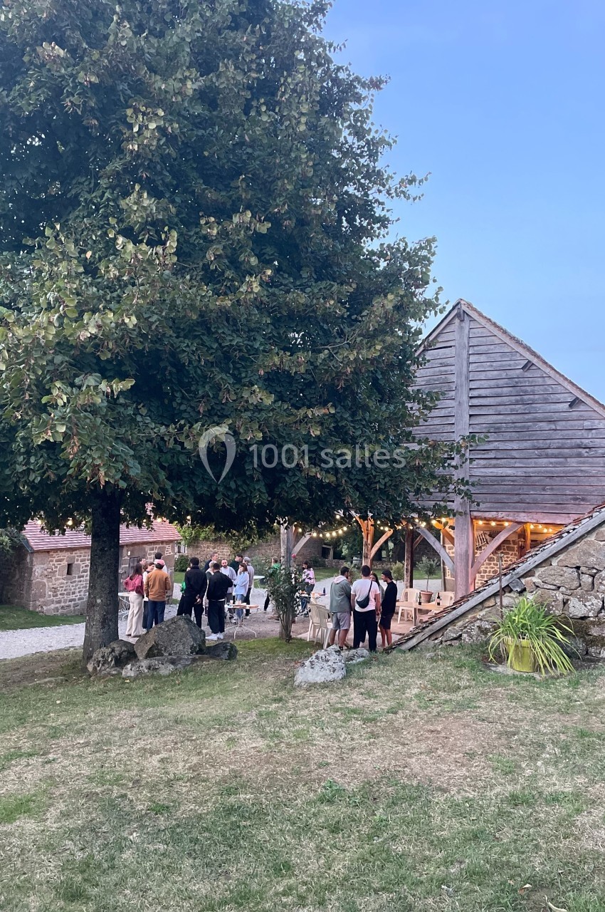 Groupe de personnes rassemblées près d'une grange en bois éclairée, entourée d'herbe et d'un grand arbre.
