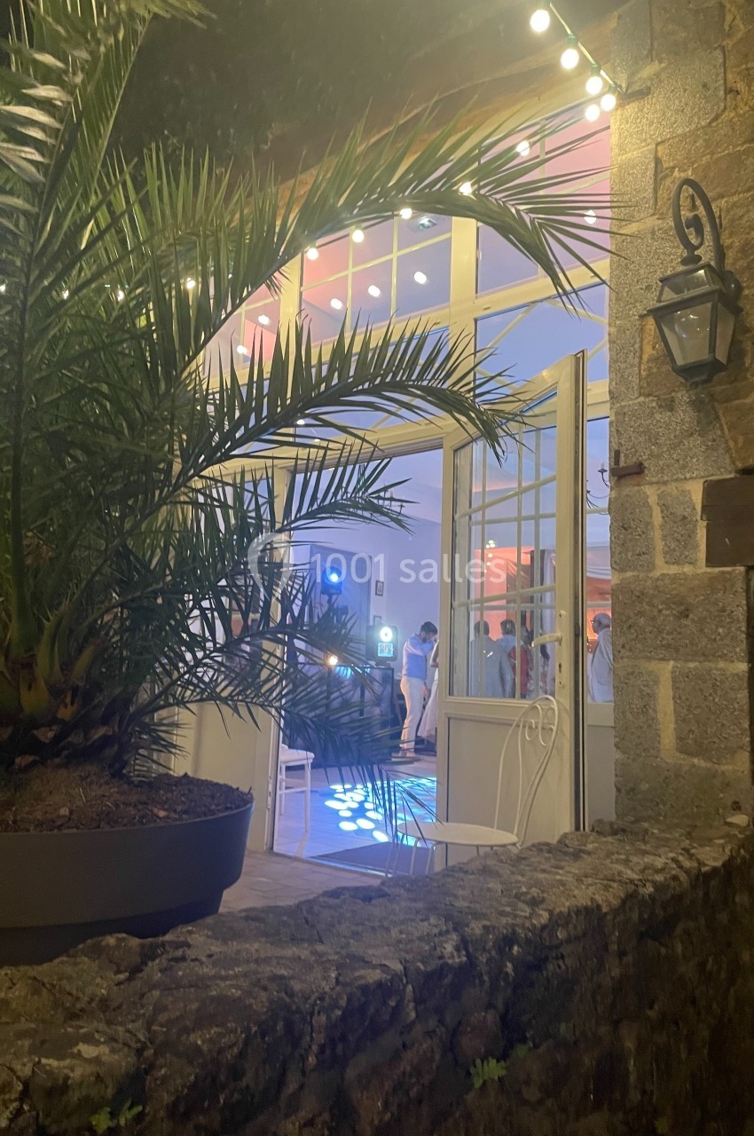 Vue nocturne d'une salle éclairée avec des invités dansant, encadrée par un palmier et un mur en pierre.