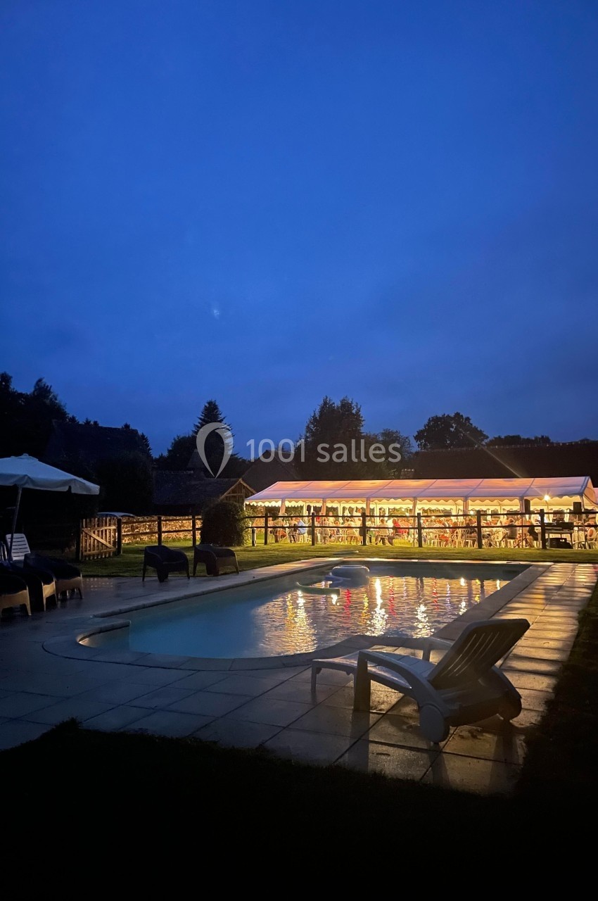 Piscine éclairée au crépuscule avec des chaises longues et un bâtiment illuminé en arrière-plan.