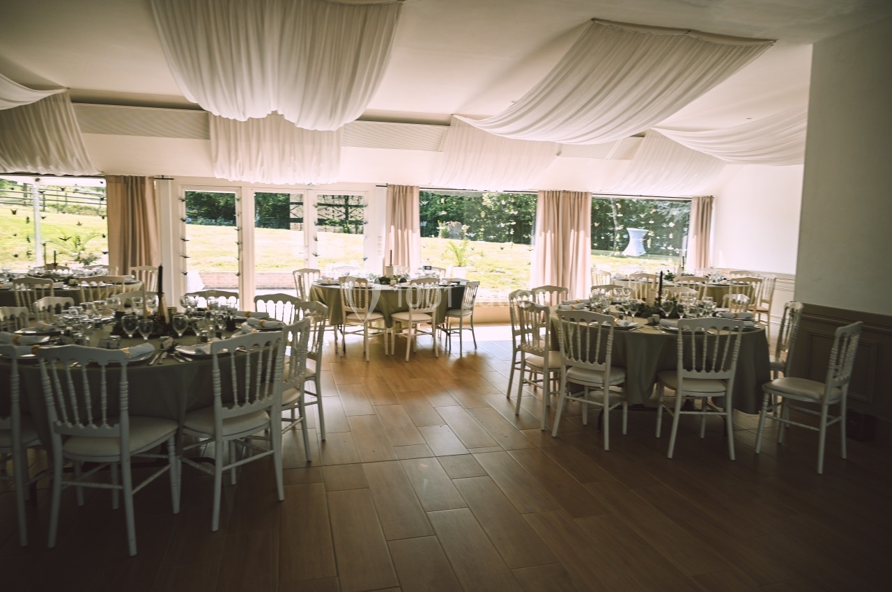 Salle de réception lumineuse avec tables rondes dressées, chaises blanches et grandes baies vitrées donnant sur un jardin.