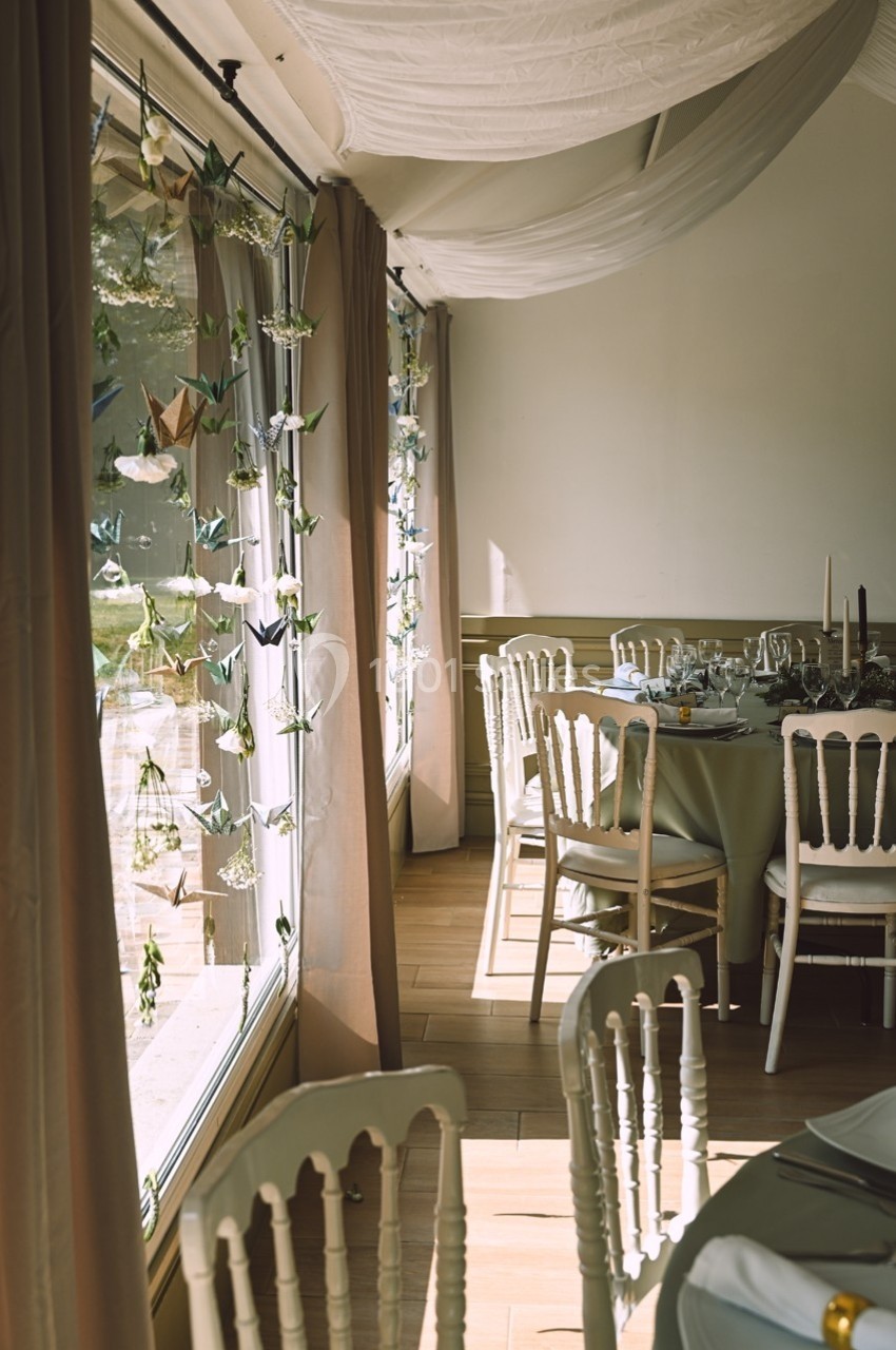 Salle de réception lumineuse avec tables dressées, chaises blanches et décorations suspendues près des fenêtres.