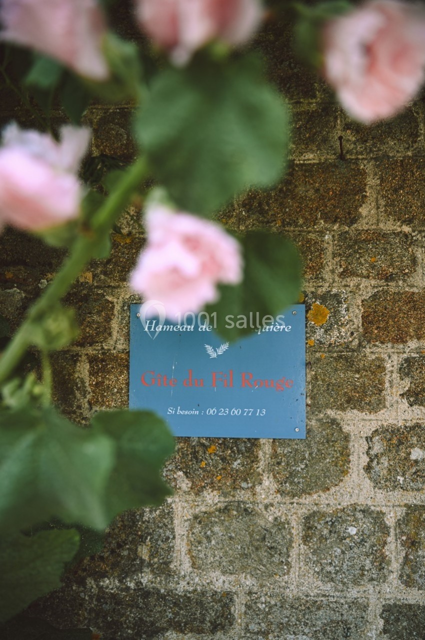 Plaque bleue indiquant ’Gîte du Fil Rouge’ fixée sur un mur en pierre, partiellement masquée par des fleurs roses.