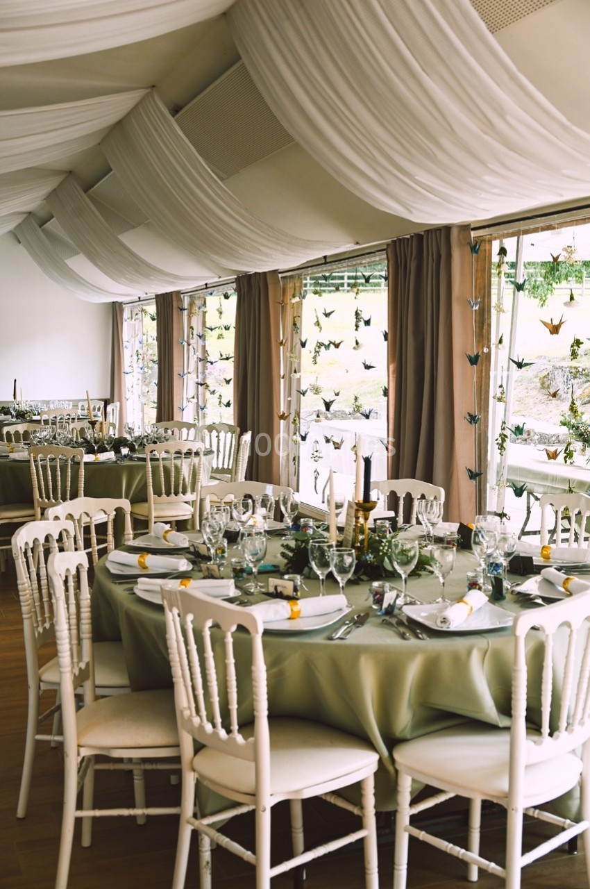 Salle de réception décorée avec des tables rondes, nappes vertes, chaises blanches et guirlandes suspendues.