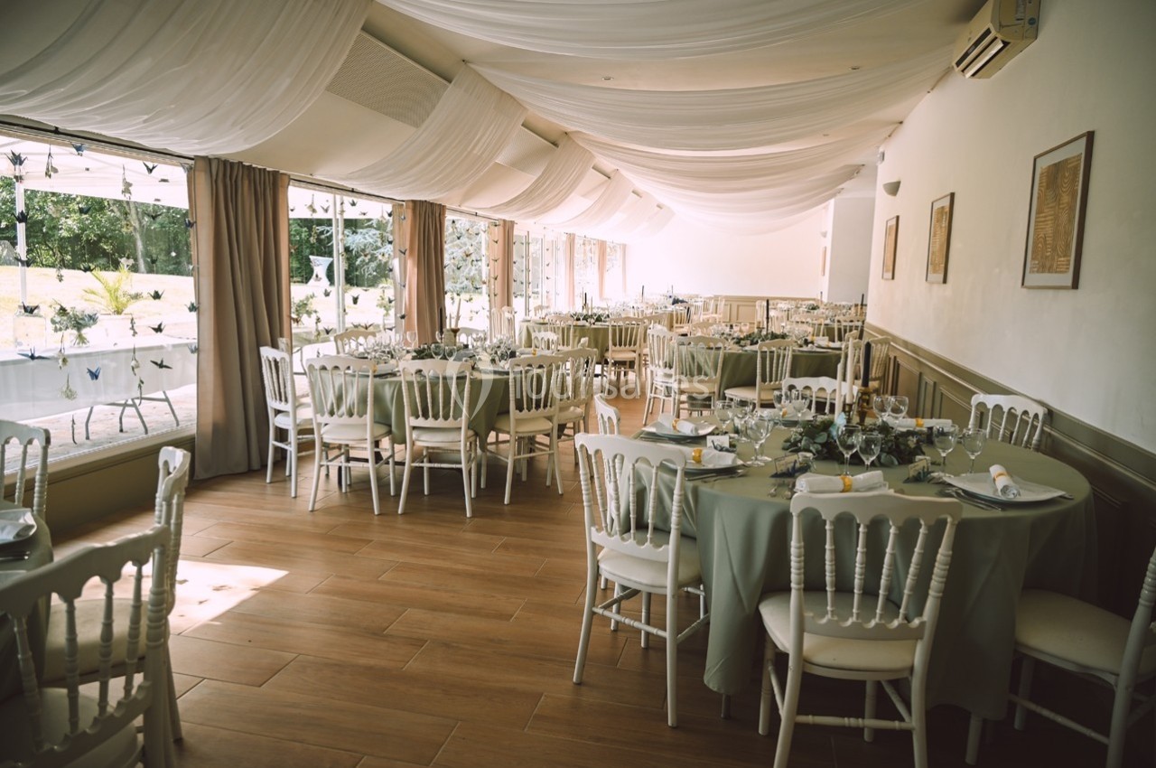 Salle de réception lumineuse avec tables rondes dressées, nappes vertes et chaises blanches, décorée de voilages au plafond.