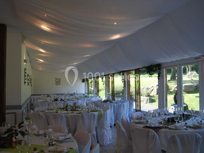 Salle de réception décorée avec des tables dressées, des nappes blanches et un plafond drapé, donnant sur un jardin.