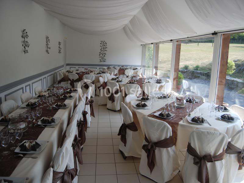 Salle de réception décorée avec des tables dressées, nappes blanches et chemins de table marron, près de grandes baies…