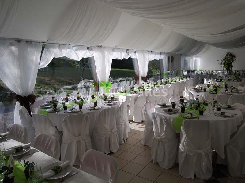 Salle de réception décorée avec des nappes blanches, des rubans verts et des tables dressées près de grandes baies vitrées.