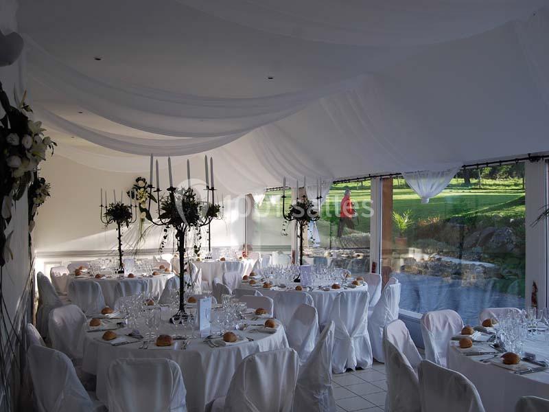 Salle de réception décorée avec des tables rondes, nappes blanches, chandeliers et vue sur un jardin à travers de grandes…