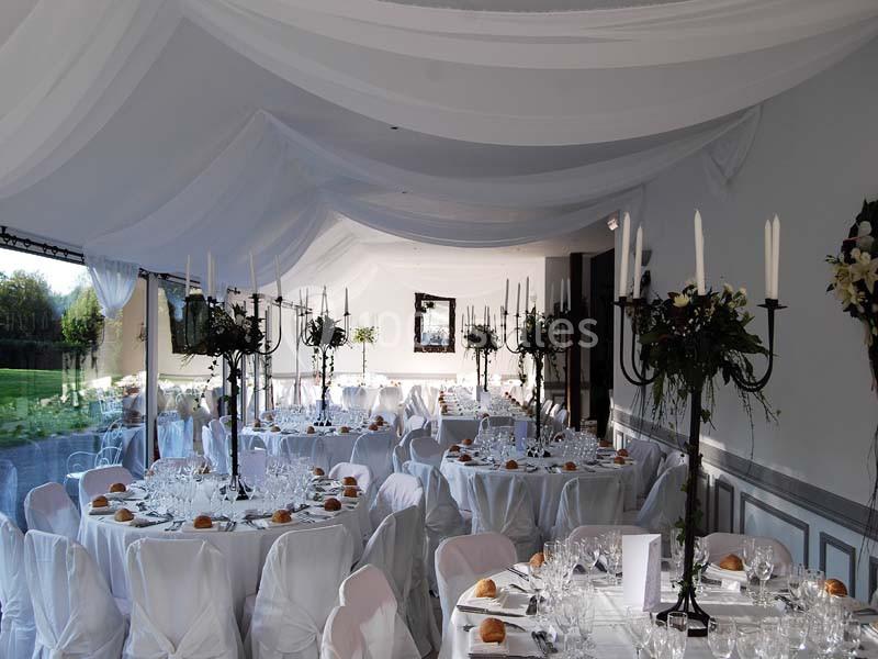 Salle de réception décorée avec des tables rondes dressées, nappes blanches, chandeliers et drapés au plafond.