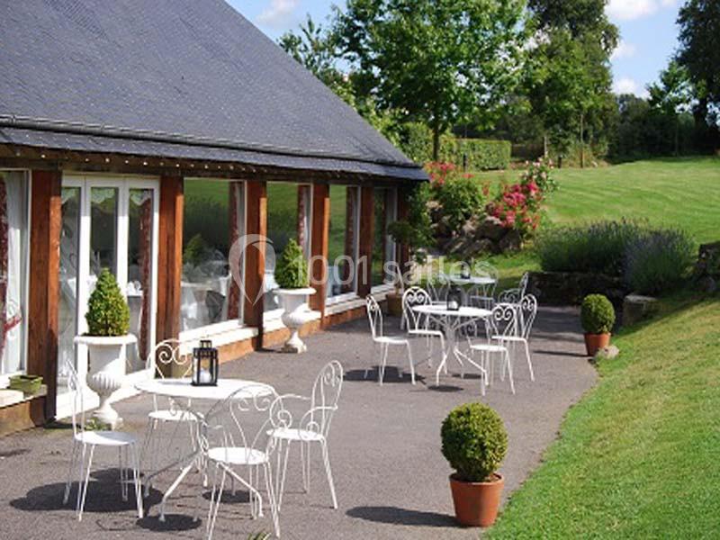 Terrasse avec tables et chaises en fer forgé, bordée de buissons et donnant sur un jardin verdoyant par temps ensoleillé.