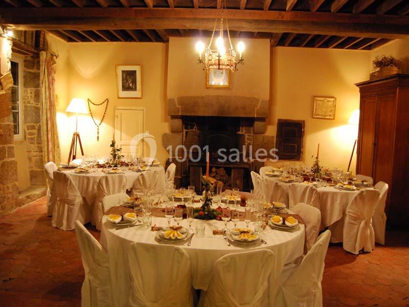 Salle de réception chaleureuse avec tables rondes dressées, nappes blanches et éclairage tamisé près d'une cheminée.