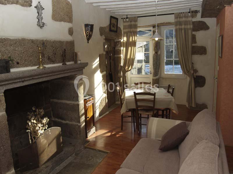 Salon rustique avec cheminée en pierre, table à manger près d'une fenêtre, canapé beige et poutres apparentes.