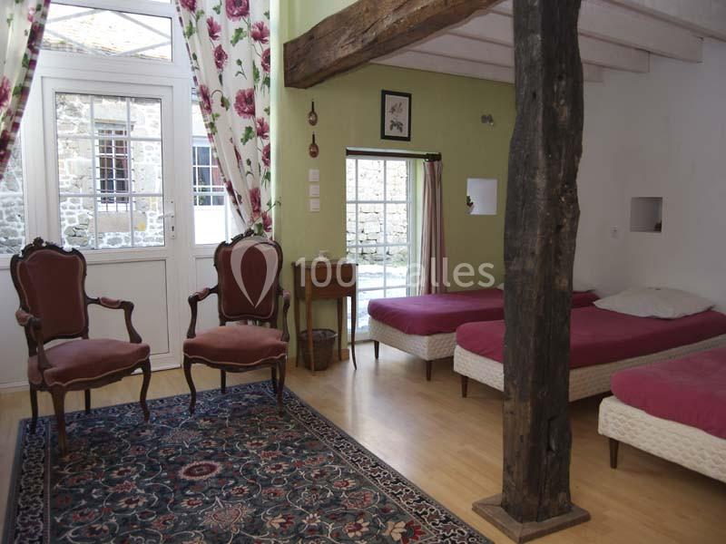 Chambre lumineuse avec lits simples, fauteuils anciens, poutre apparente, tapis et vue sur une cour pavée.