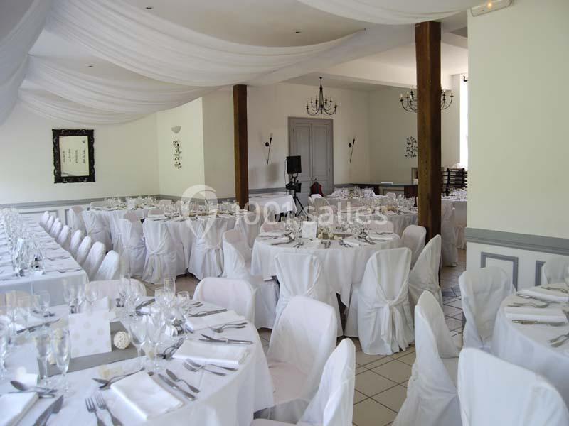 Salle de réception décorée avec des tables rondes et rectangulaires dressées, chaises couvertes de housses blanches.