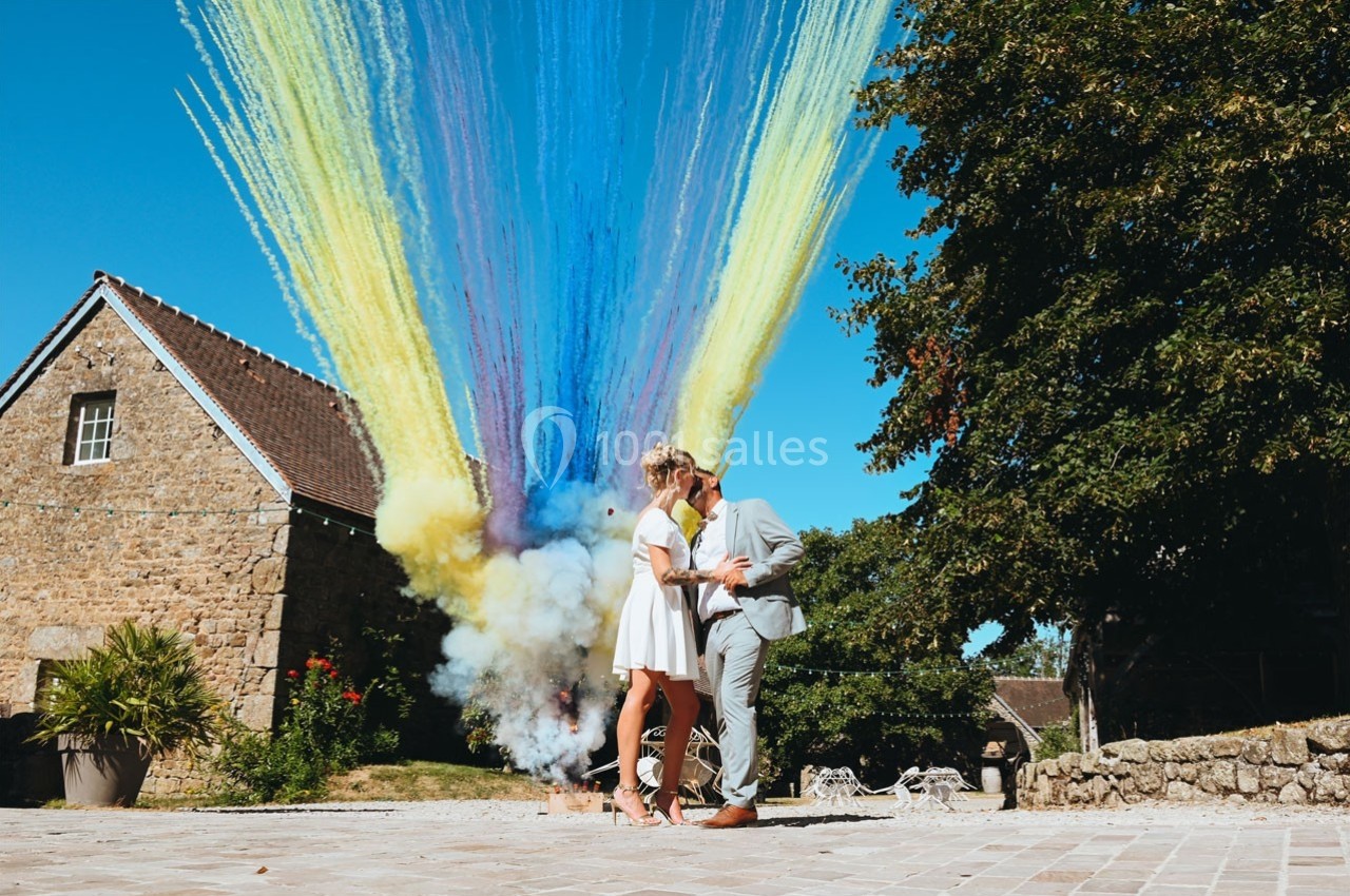 Un couple s'embrasse devant une maison en pierre, avec des fumées colorées jaillissant en arrière-plan.