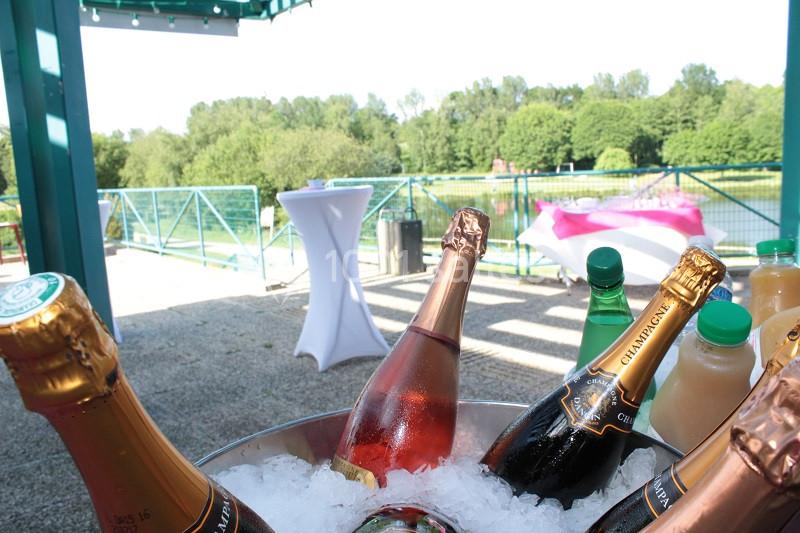 Bouteilles de champagne et de jus dans un seau de glace, avec vue sur un lac et une terrasse en arrière-plan.