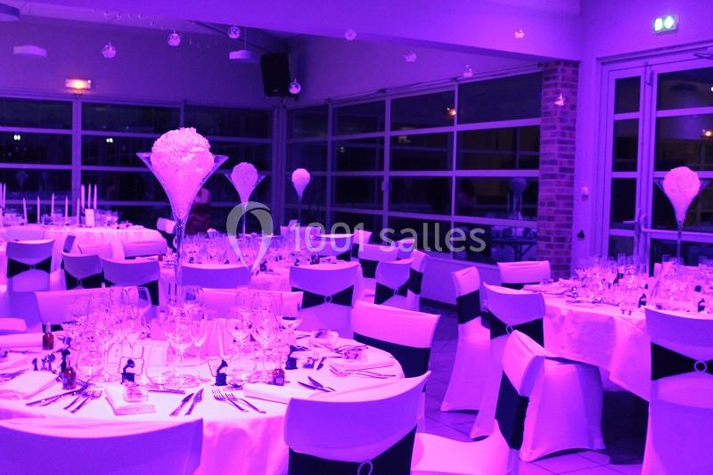 Salle de réception décorée avec des tables rondes, nappes blanches, chaises ornées et éclairage violet tamisé.