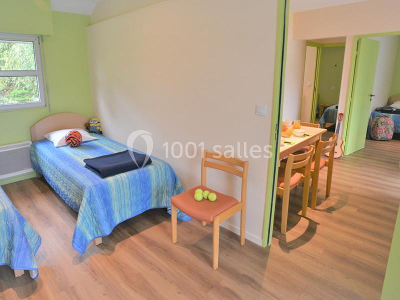 Chambre avec deux lits simples, sol en parquet, chaise avec balles de tennis, table et porte ouverte sur une autre pièce.