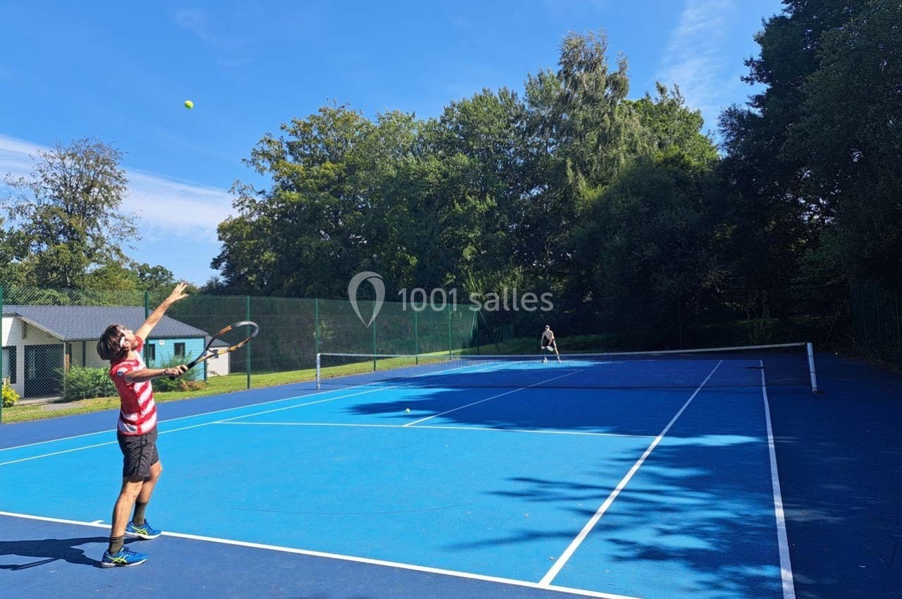 Un joueur de tennis effectue un service sur un court extérieur entouré d'arbres par temps ensoleillé.