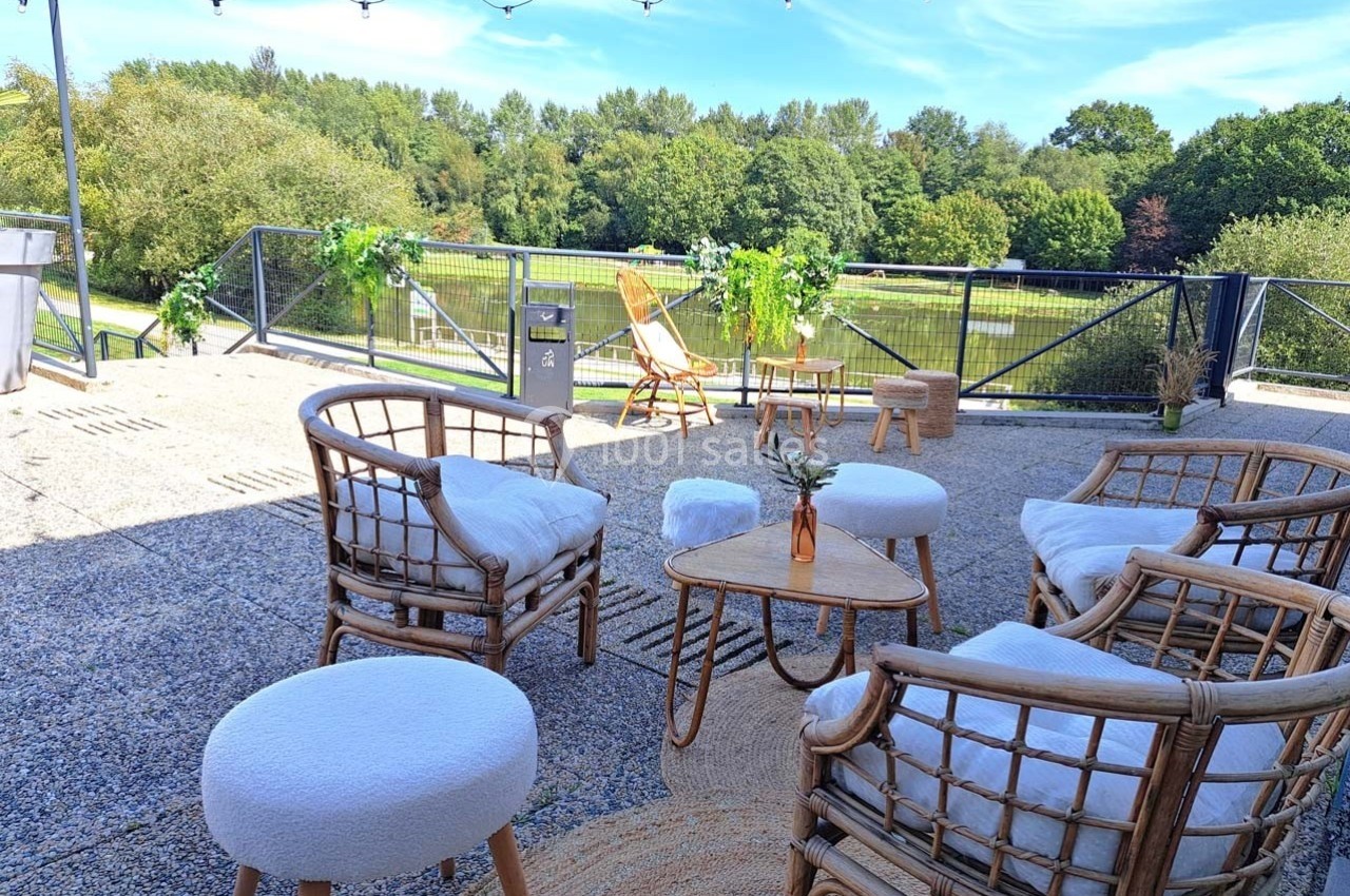 Terrasse aménagée avec fauteuils en rotin, table basse et vue sur un étang entouré de verdure par une journée ensoleillée.