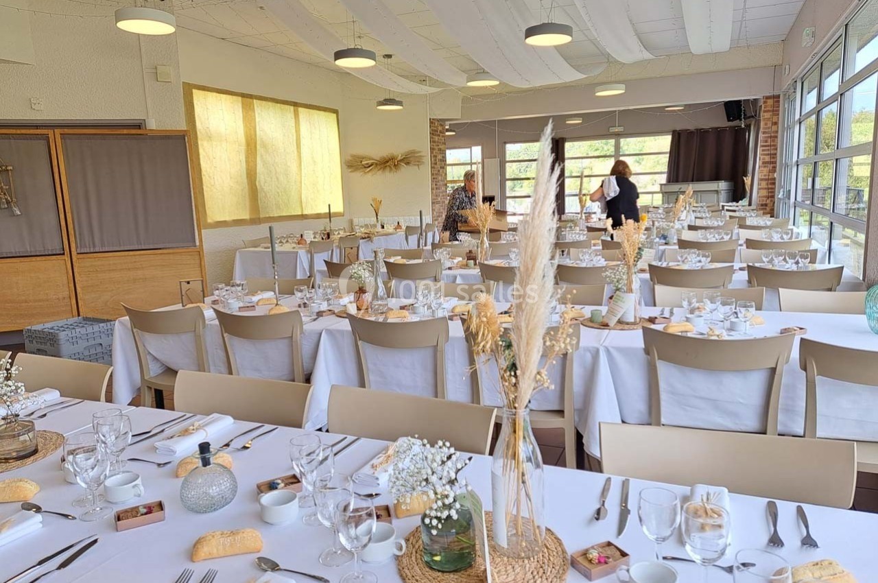 Salle de réception lumineuse avec tables dressées, nappes blanches, décorations florales et grandes baies vitrées.