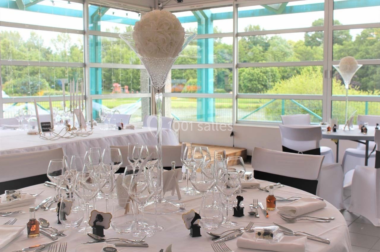 Salle de réception lumineuse avec tables dressées, nappes blanches, verres, et centre de table floral en hauteur.