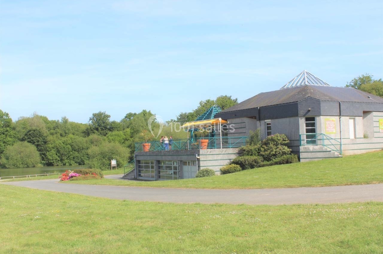 Bâtiment moderne avec terrasse surélevée, entouré de verdure et situé près d'un plan d'eau.