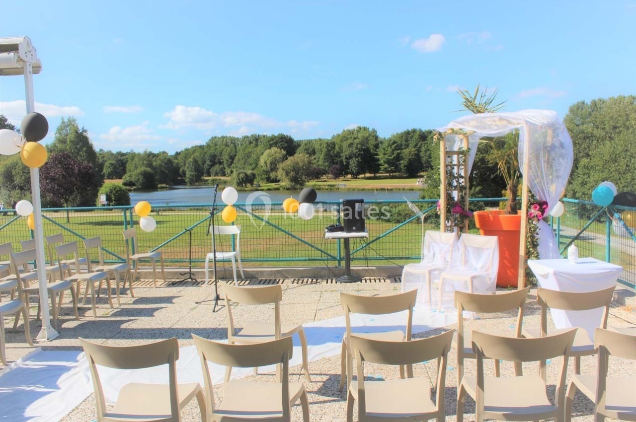 Chaises disposées face à une arche décorée, avec vue sur un lac et un paysage verdoyant sous un ciel bleu.