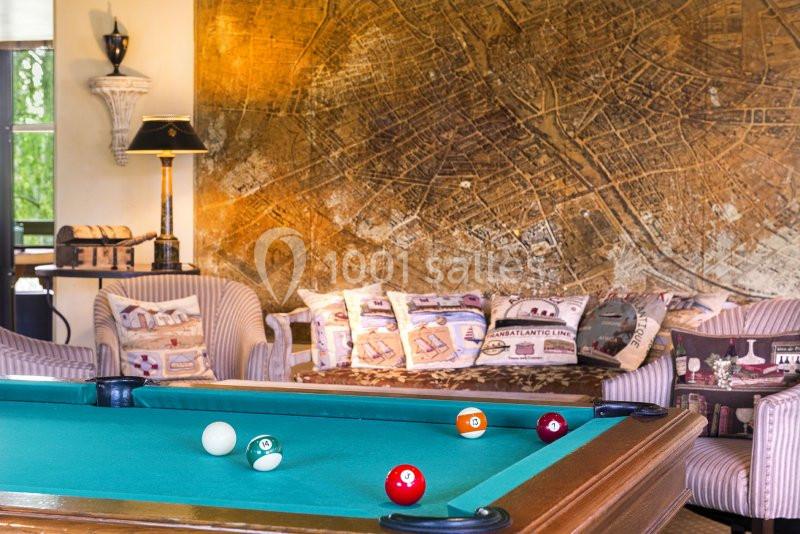 Table de billard avec des boules colorées au premier plan, entourée de fauteuils et d'une carte murale en arrière-plan.