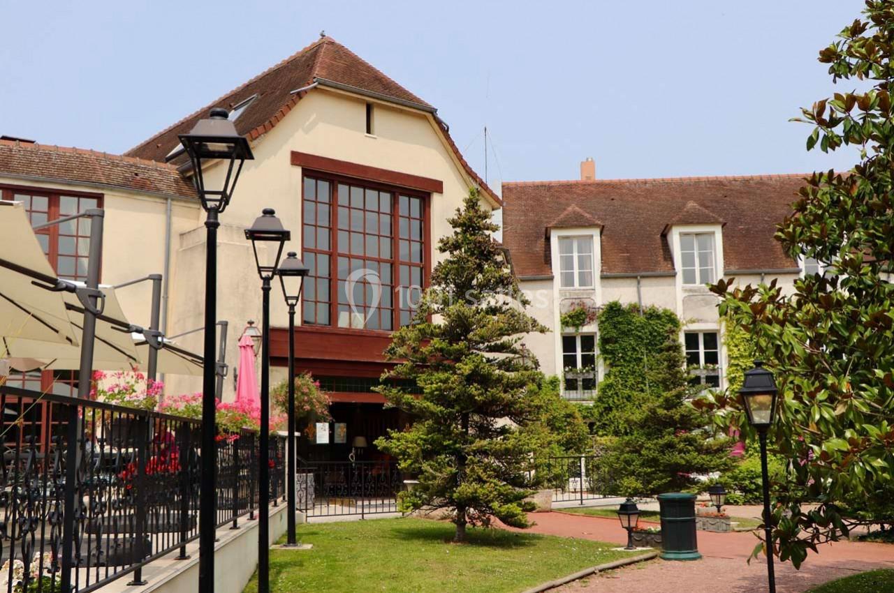 Façade d'un bâtiment avec grandes fenêtres, terrasse ombragée, lampadaires et jardin arboré au premier plan.