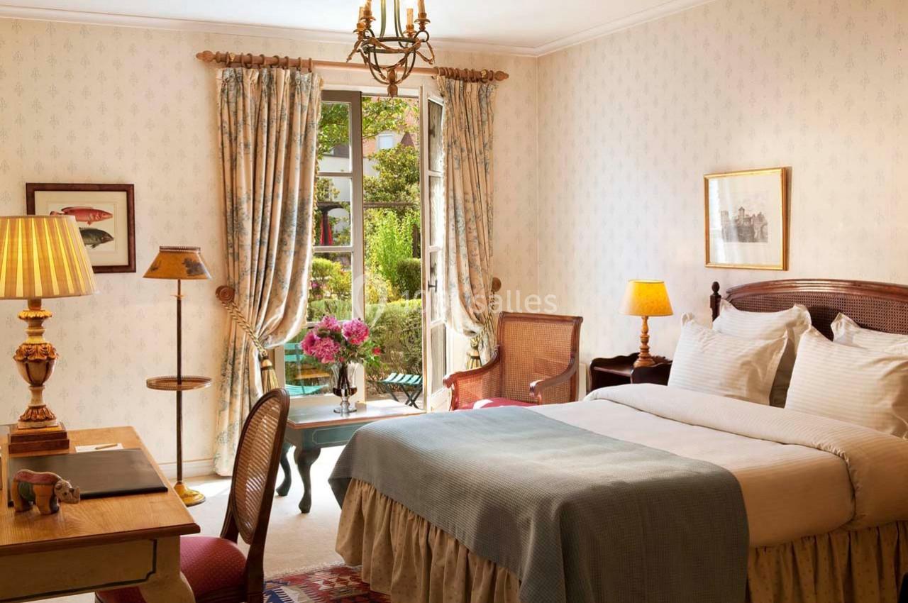 Chambre lumineuse avec lit double, bureau en bois, fauteuils et vue sur un jardin à travers une porte-fenêtre ouverte.