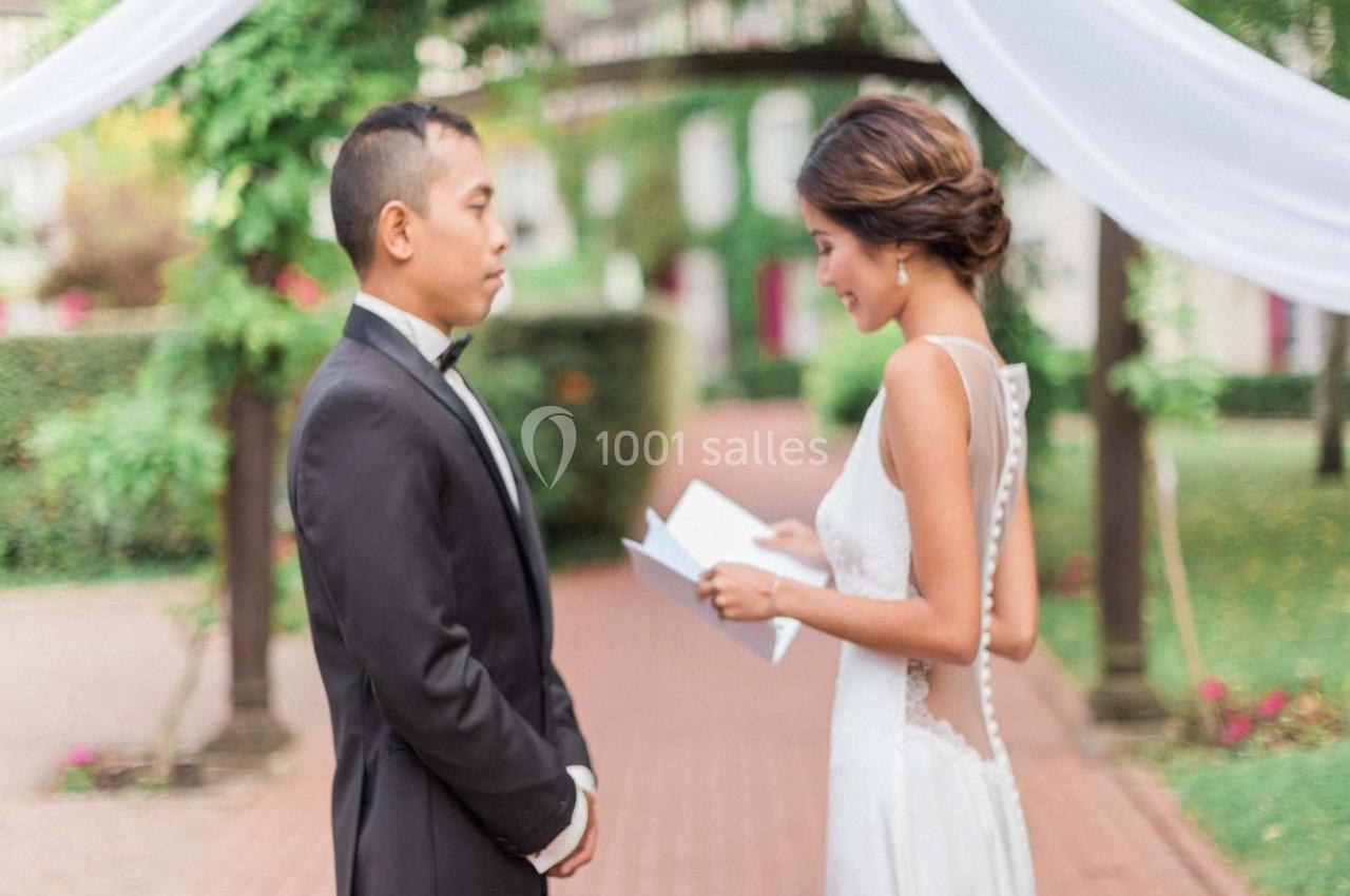Une femme en robe de mariée lit un texte devant un homme en costume dans un jardin décoré de voilages blancs.