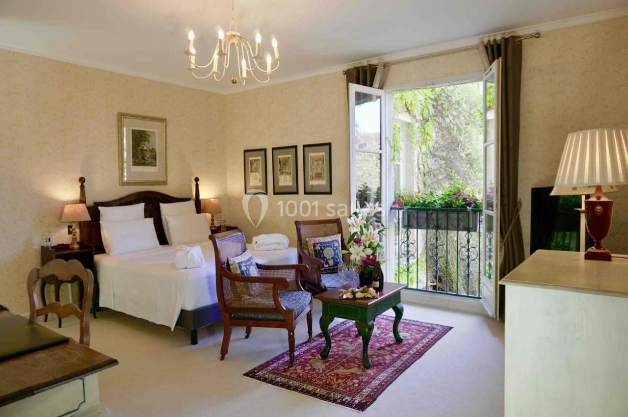 Chambre lumineuse avec lit double, coin salon, balcon ouvert sur un jardin et décoration classique.