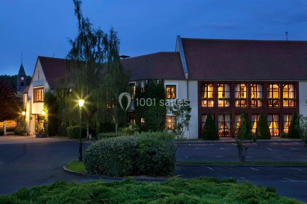 Façade d'un bâtiment éclairé à la tombée de la nuit, entouré de végétation et d'un parking partiellement visible.