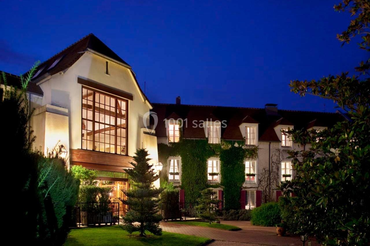 Façade d'un bâtiment éclairé de nuit, entouré de végétation et d'un chemin menant à l'entrée principale.