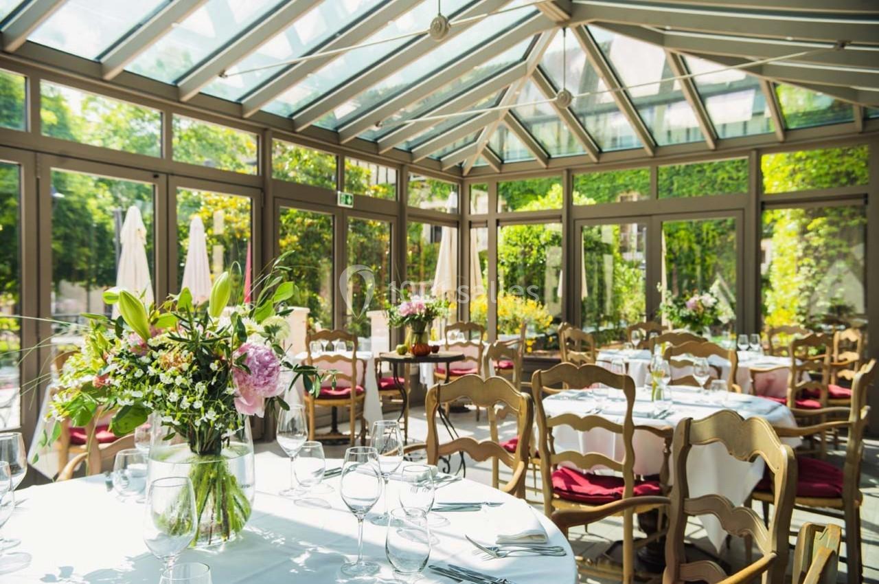Salle lumineuse avec verrière, tables dressées avec nappes blanches, chaises en bois et bouquets de fleurs.