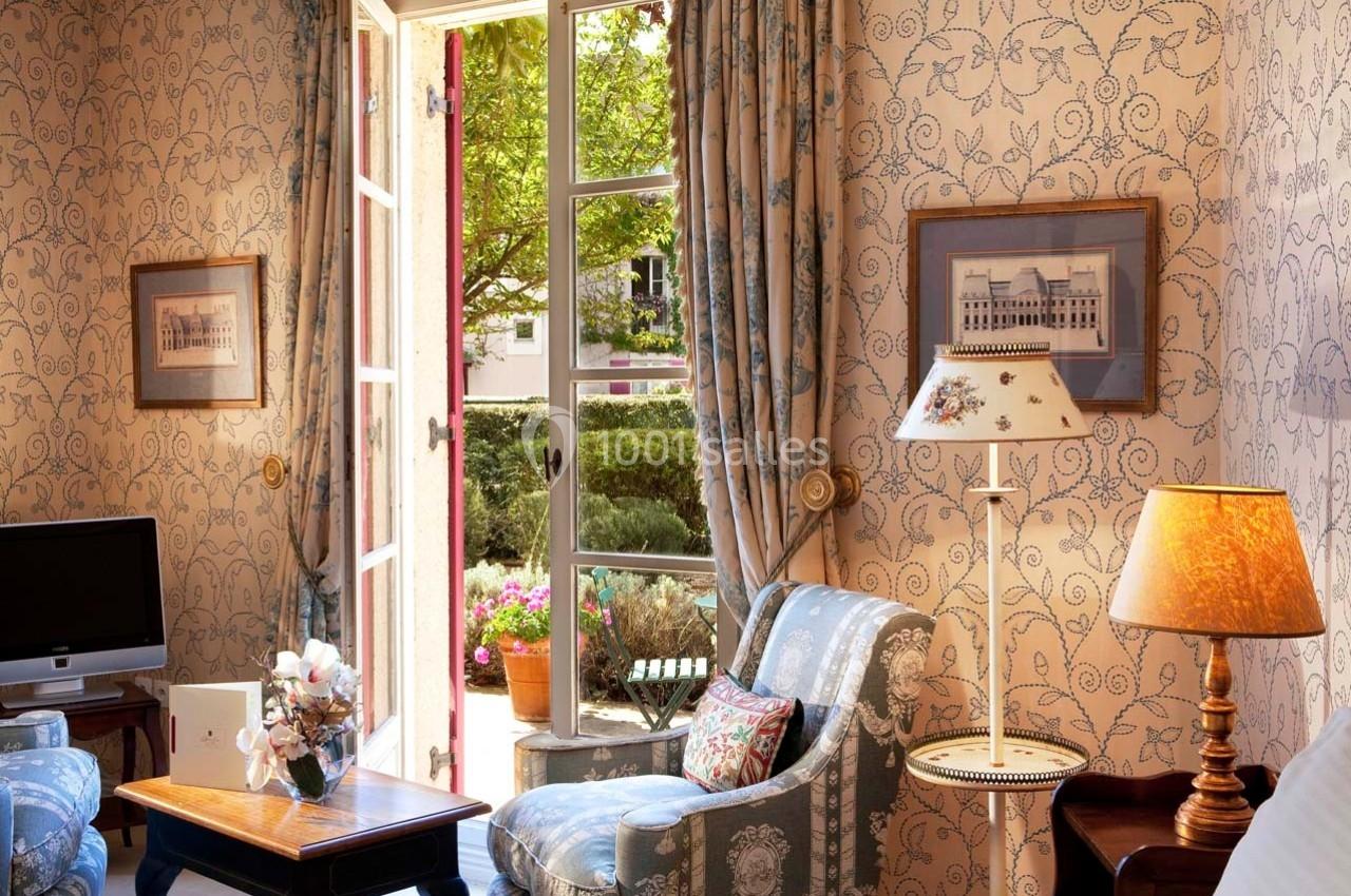 Salon lumineux avec fauteuils, table basse, lampe et fenêtre ouverte donnant sur un jardin verdoyant.