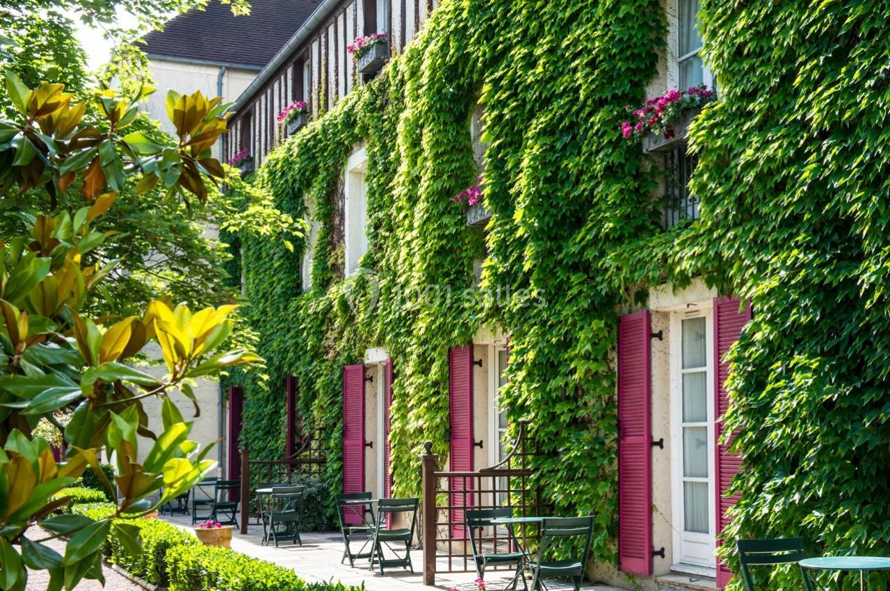 Façade d'un bâtiment recouverte de lierre avec volets roses, terrasses aménagées et végétation environnante.