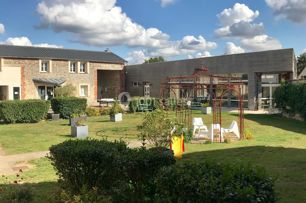 Jardin aménagé avec des arbustes, une tonnelle en métal, des chaises blanches et des bâtiments en arrière-plan.