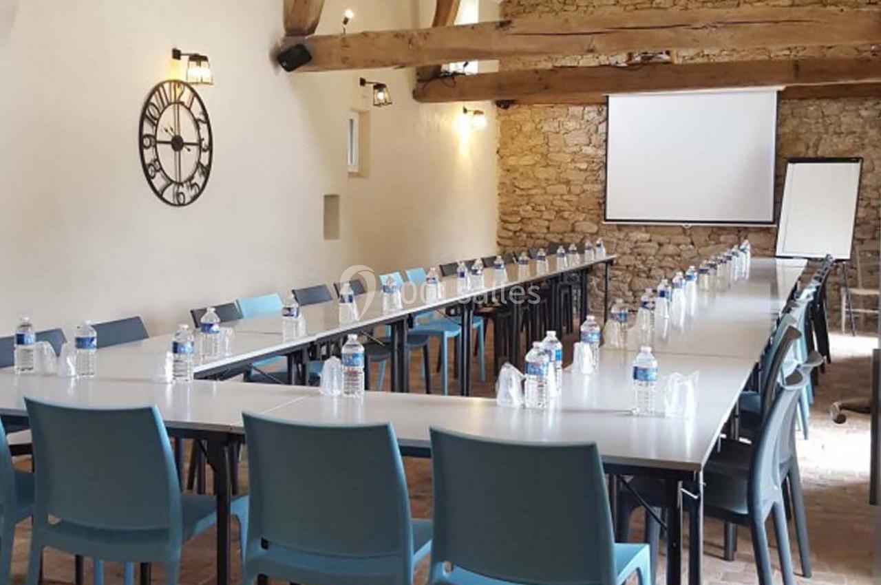 Salle de réunion avec tables en U, chaises bleues, bouteilles d'eau, mur en pierre, écran de projection et tableau blanc.