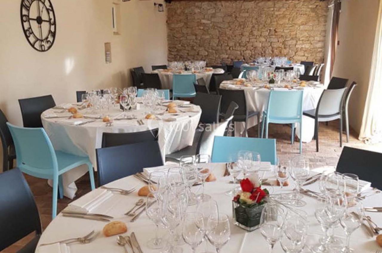 Salle de réception avec des tables rondes dressées, nappes blanches, chaises modernes et mur en pierre apparente.