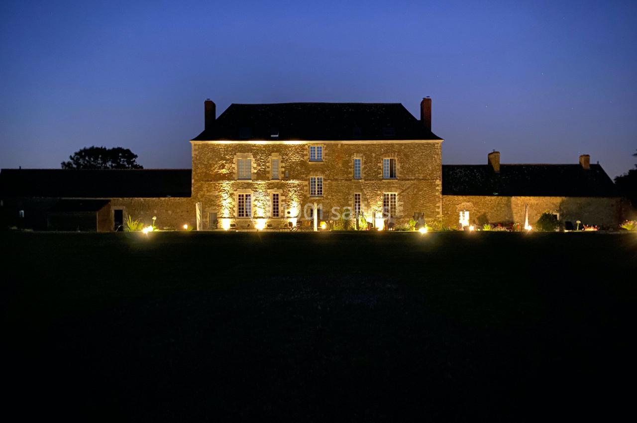 Façade d'une grande maison en pierre éclairée la nuit, entourée d'un jardin sombre et d'annexes.