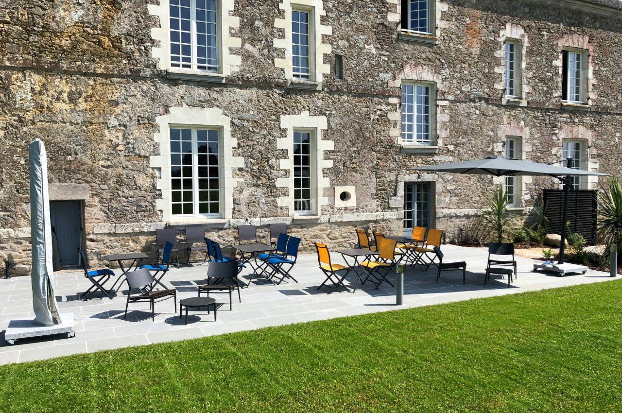 Terrasse en pierre avec tables, chaises colorées et parasol, devant une façade en pierre avec grandes fenêtres.